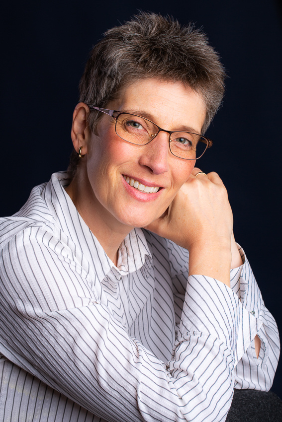 Sabine M. Schröder schaut fröhlich lächelnd schräg in die Kamera. Ihr Kopf ist leicht auf die Hände gestützt, sie trägt eine weiße Bluse mit grauen dünnen Streifen.